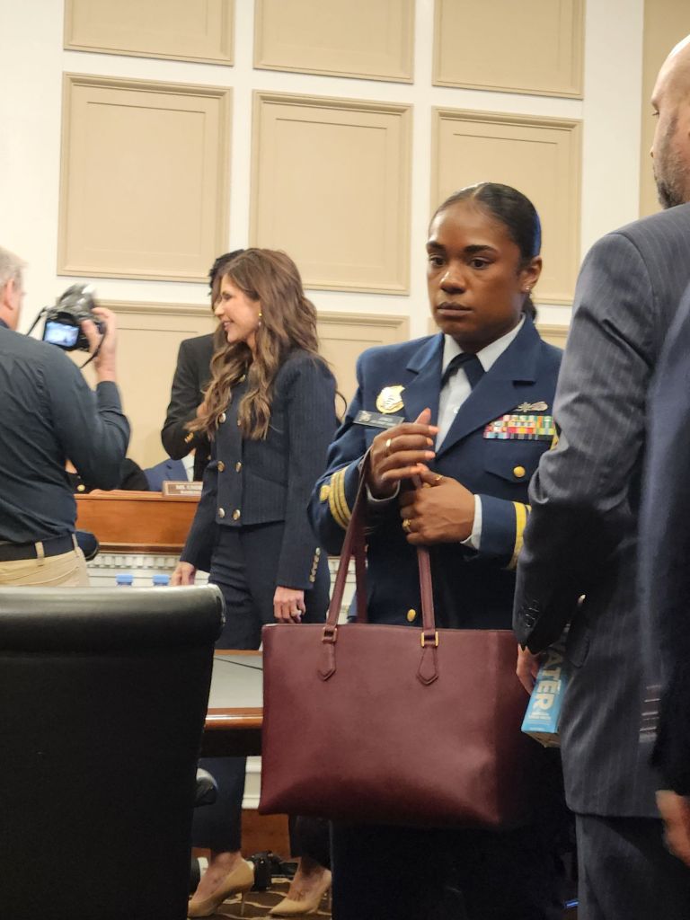 A military officer in uniform holding a burgundy handbag, standing among a group of people in a formal setting.