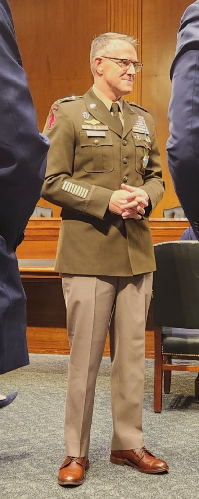 A military officer in uniform standing in a formal setting, with various medals and insignia displayed on his uniform.