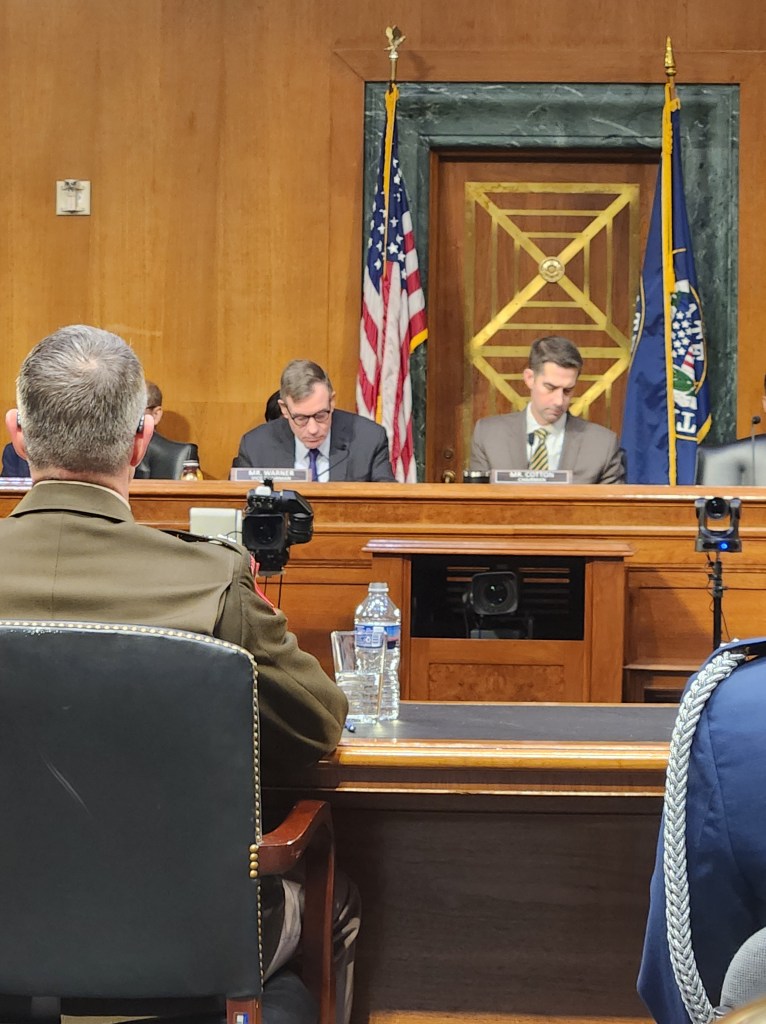 A view of a formal meeting with several individuals seated at a table, including two attendees focused on their notes. American flags are displayed in the background.